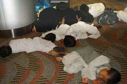 Out cold. Five small boys asleep on the floor of the Dubai airport. An older boy sits off to the right. A girl cuddles against her sleeping mother, in black. Somewhere, there must be an infant, as a baby bottle is standing behind the mother. Eventually the father arrived and squatted over his brood.